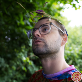 A white masc human, Laurie, surrounded by greenery in the blurry background, gazing into the middle distance. They are wearing a wreath of flowers on their head and large glasses with transparent frames.