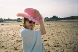 Photo of a person standing on a beach. They are a white femme presenting person, in the middle of the frame, wearing a pink cowboy hat and sunglasses, wrapped in a towel. Their hand is on the top of their hat like they are about to cock it in a sort of ‘howdy’ way. Behind them is sand and a line of trees on the horizon.